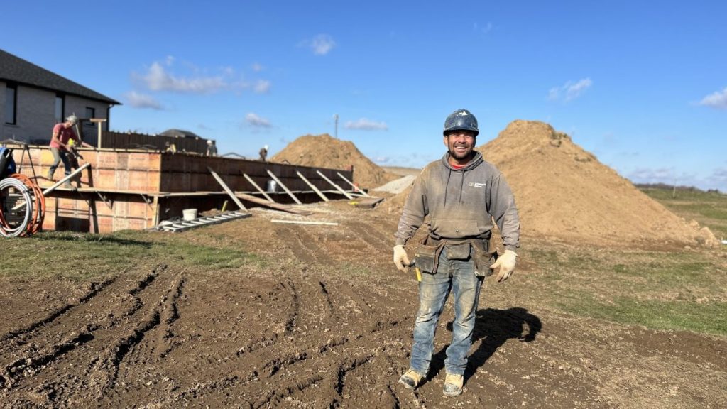 A construction worker in a helmet and work clothes stands in front of a foundation structure and dirt mounds at a building site on a clear day.