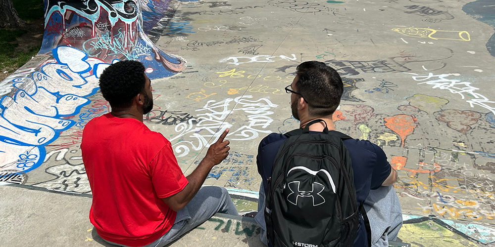 A male youth worker and male client sit on the edge of a graffiti-covered skate park; the worker wears a red shirt and gestures while talking, and the youth wears glasses and a black backpack, listening attentively.