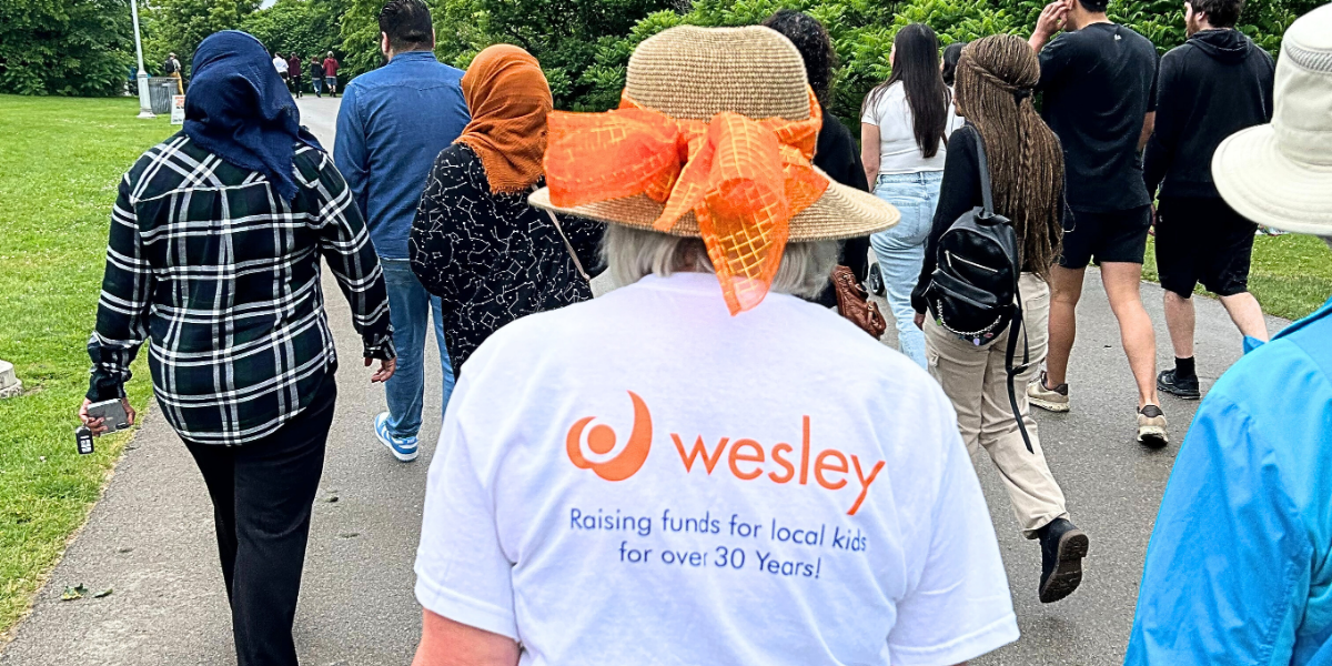 A group of people walk outdoors on a paved path. One person in front wears a straw hat with an orange bow and a shirt reading Wesley: Raising funds for local kids for over 30 years!