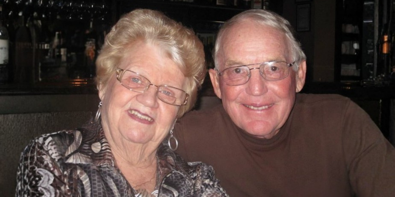 An older couple smiling together indoors, both wearing glasses. The woman has short light hair and a patterned top, and the man has short gray hair and a brown shirt.