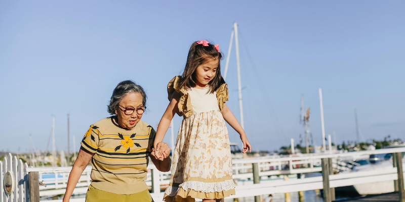 An elderly woman and a young girl holding hands while walking near a marina on a sunny day.