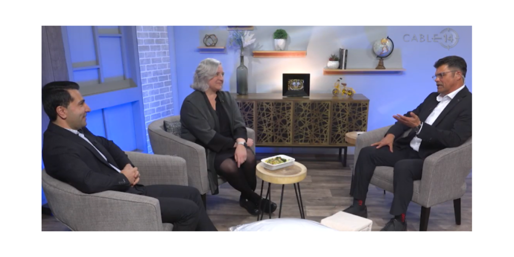 Three people seated in a TV studio during a Cable 14 Hamilton Life interview, with the host speaking to two Wesley representatives; a small table with a plated meal sits between them.