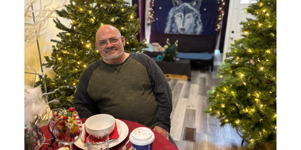 Michael sits at a table in his apartment, smiling gently, with decorated Christmas trees glowing on either side of him, reflecting a warm, festive, and welcoming home environment.