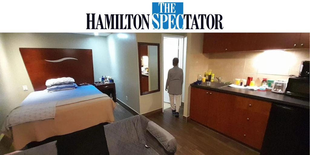 Interior photo of a small studio-style room with a made bed, kitchenette, and bathroom doorway, as a person walks toward the bathroom; the Hamilton Spectator logo appears at the top of the image.