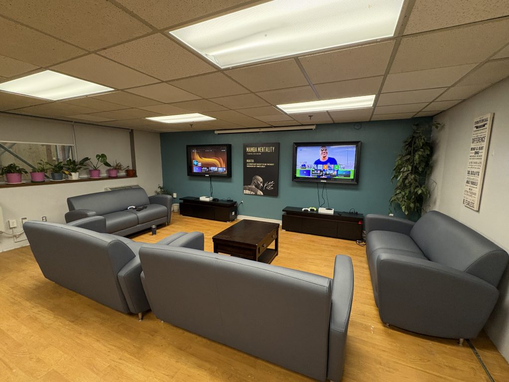 A bright, welcoming youth centre lounge with several blue couches arranged in a circle around a wooden coffee table, facing two wall-mounted TVs with gaming consoles below. Potted plants, wall art, and warm flooring create a comfortable, relaxed space for youth to gather, play games, and unwind together.