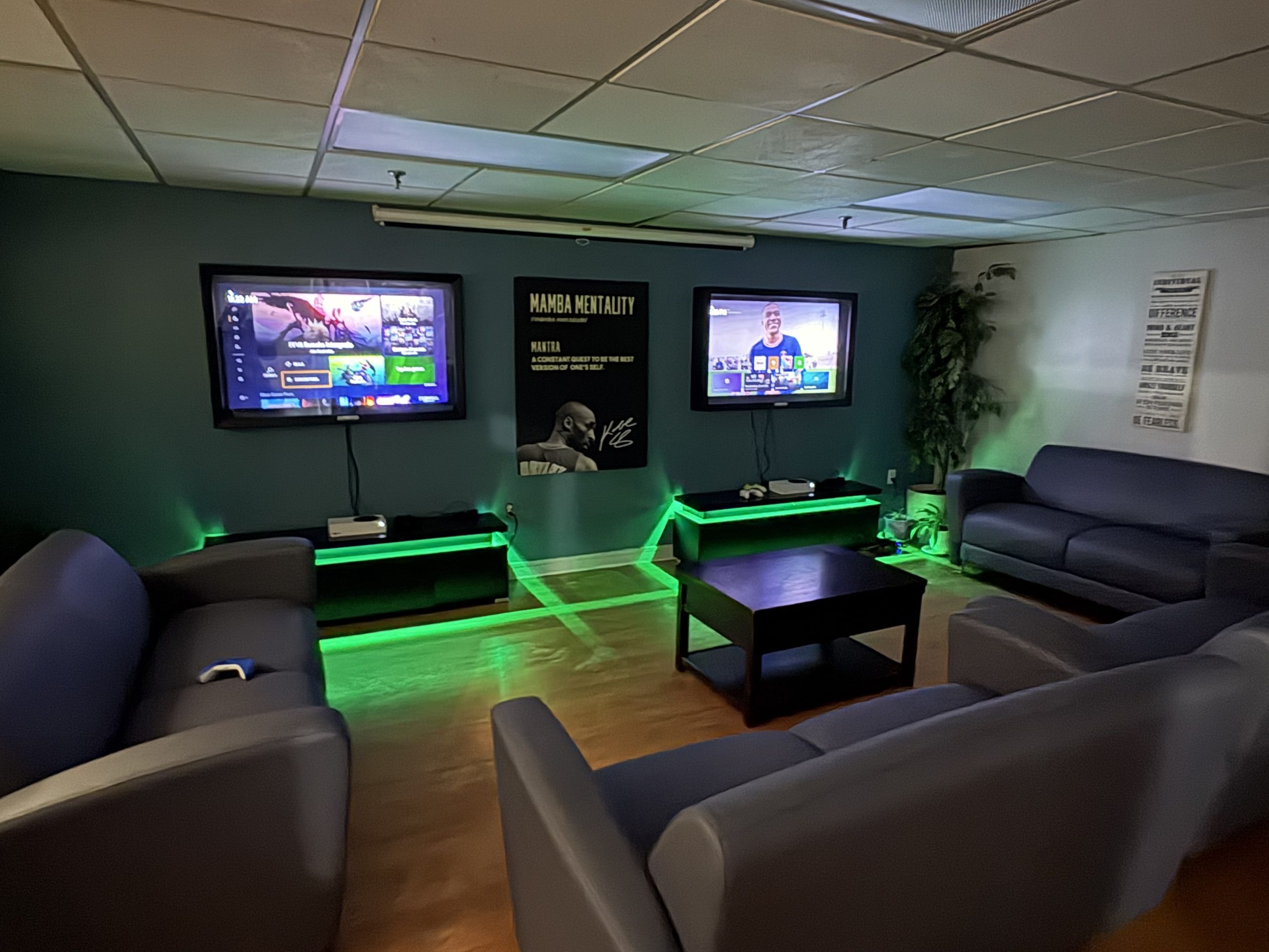 A cozy youth centre lounge with multiple blue couches arranged around a coffee table, two wall-mounted TVs displaying a video game menu, and gaming consoles below. Green LED accent lighting glows beneath the TV stands, creating a welcoming, modern space for youth to relax, play games, and socialize.