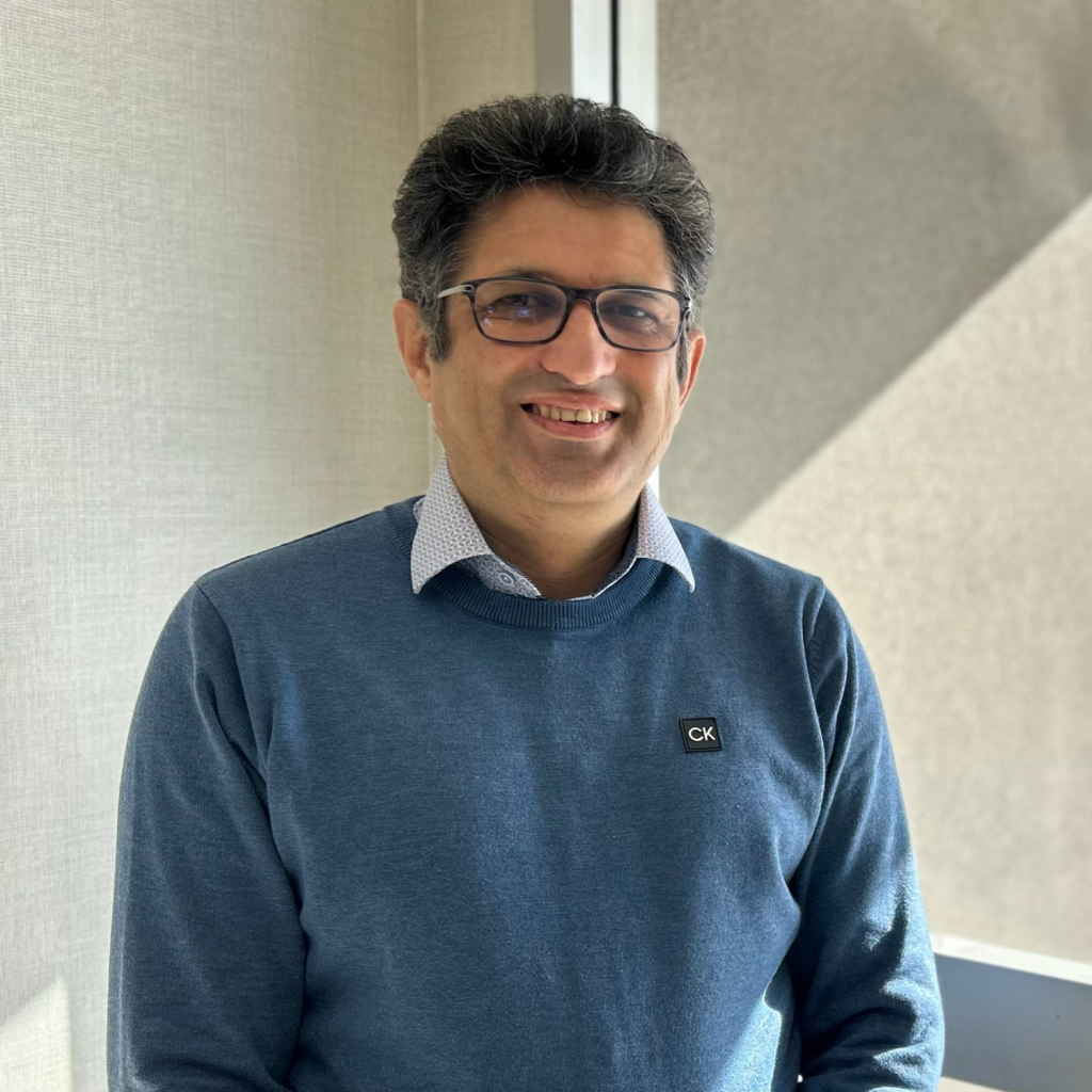 Yaqoob, wearing glasses and a blue sweater smiling indoors near a window.