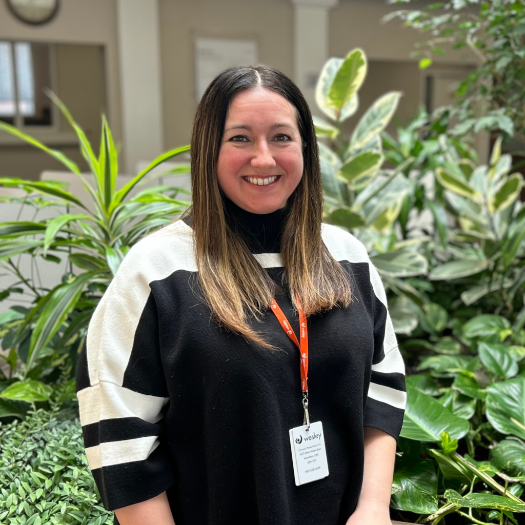 Keira, with long brown hair, wearing a black and white striped sweater and a Wesley name badge, stands and smiles in front of green plants indoors.