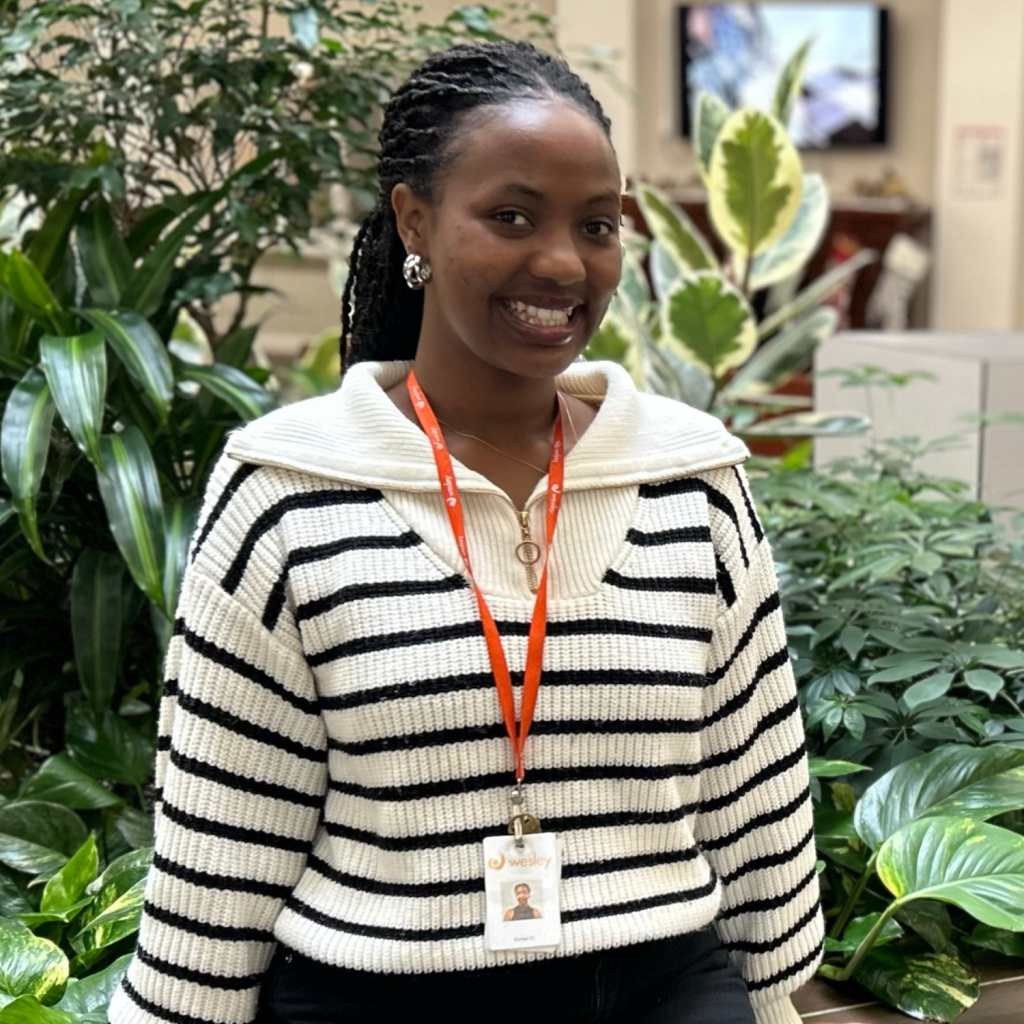 A woman wearing a striped sweater and ID badge smiles while standing next to indoor plants in an office setting.