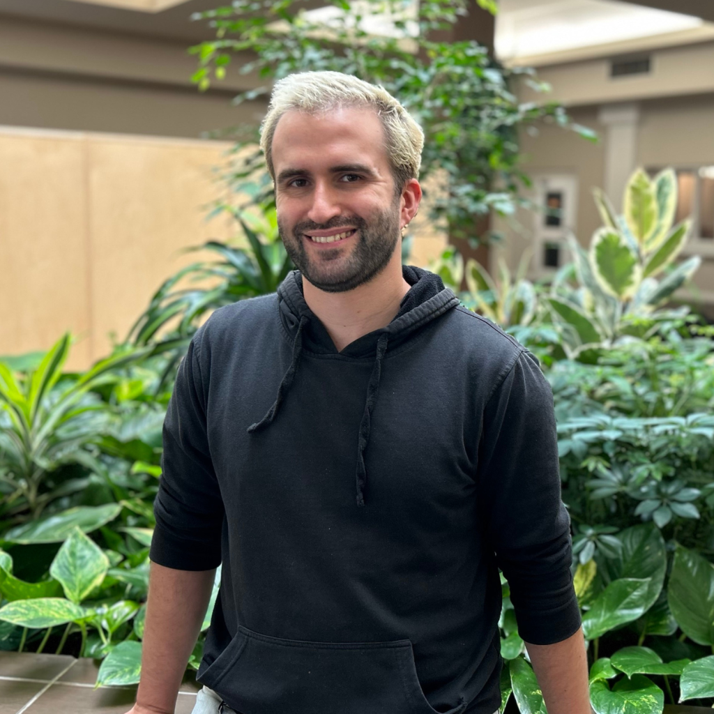 A man with short blonde hair and a dark beard, wearing a black hoodie, stands in front of indoor plants in an office setting.