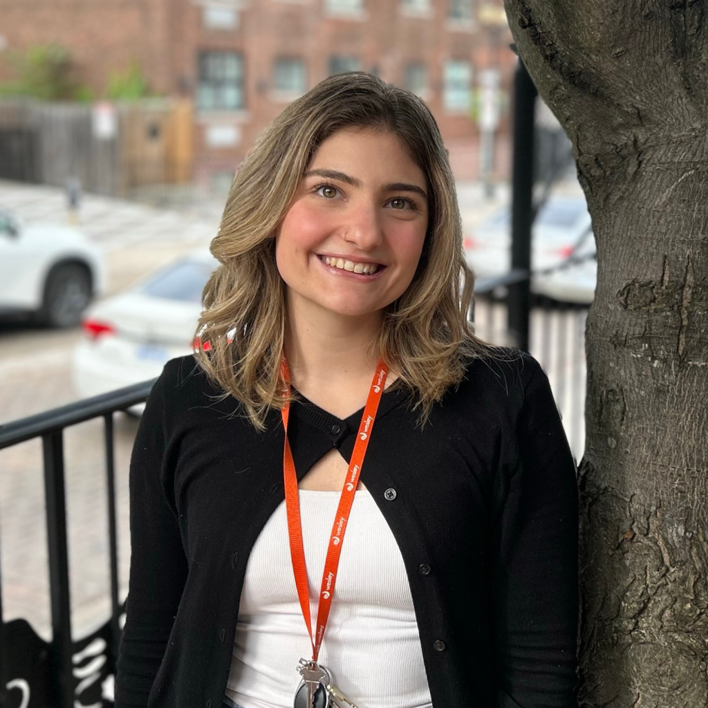 A young woman with a lanyard and keys stands next to a tree on an outdoor patio, smiling at the camera. Cars and brick buildings are visible in the background.