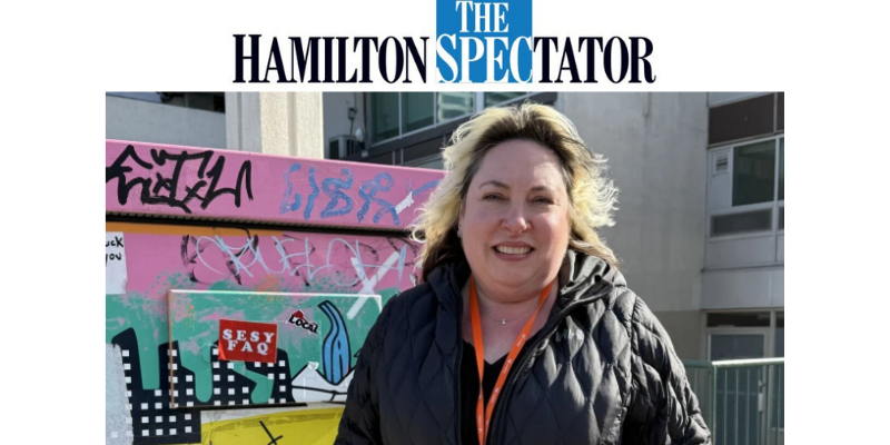 A person wearing a black quilted jacket and an orange lanyard with an ID badge stands outdoors beside a brightly coloured, graffiti-covered utility box. The artwork features bold shapes, cityscape elements, and layered street-art tags. Concrete buildings and railings appear in the background on a sunny day.