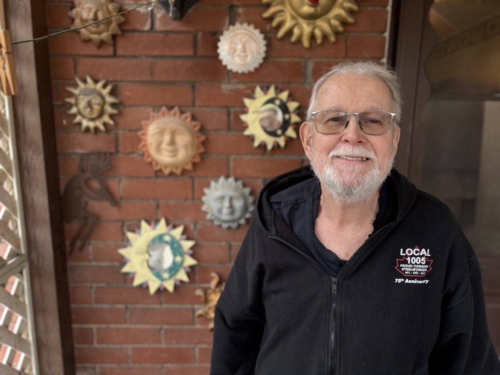 An older man with glasses and a white beard smiles at the camera, standing in front of a brick wall decorated with colourful sun face ornaments. He is wearing a black hoodie with a union logo.