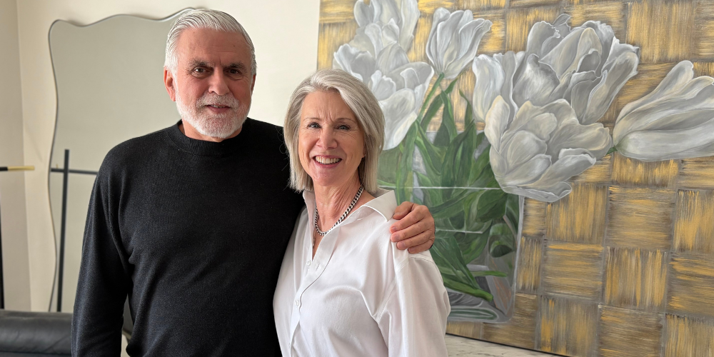 A couple stands close together indoors, smiling at the camera. The man wears a black sweater with his arm around the woman, who is dressed in a white blouse. Behind them is a large painting of white flowers against a textured gold background.