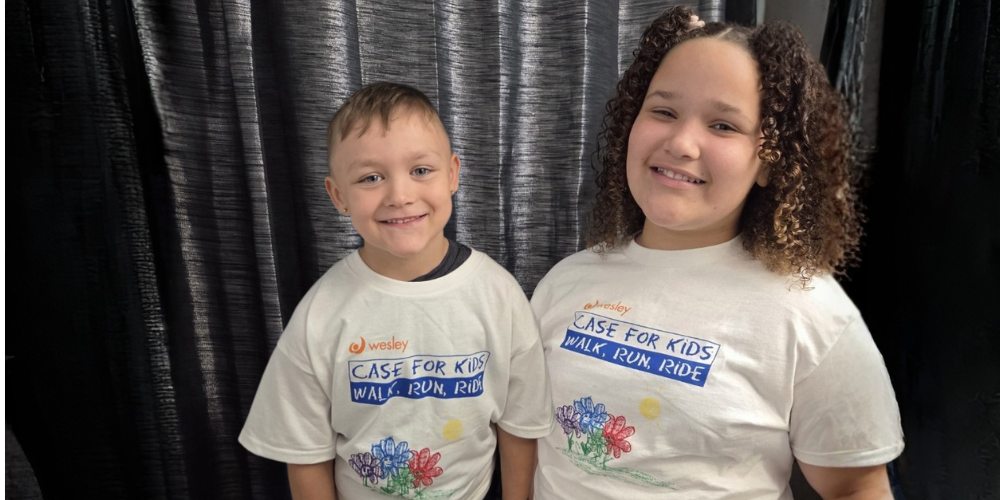 Two smiling children stand in front of a black curtain backdrop wearing white Wesley Case for Kids t-shirts that read &ldquo;Walk, Run, Ride.&rdquo; The shirts feature a colourful flower design, and both children are smiling at the camera.