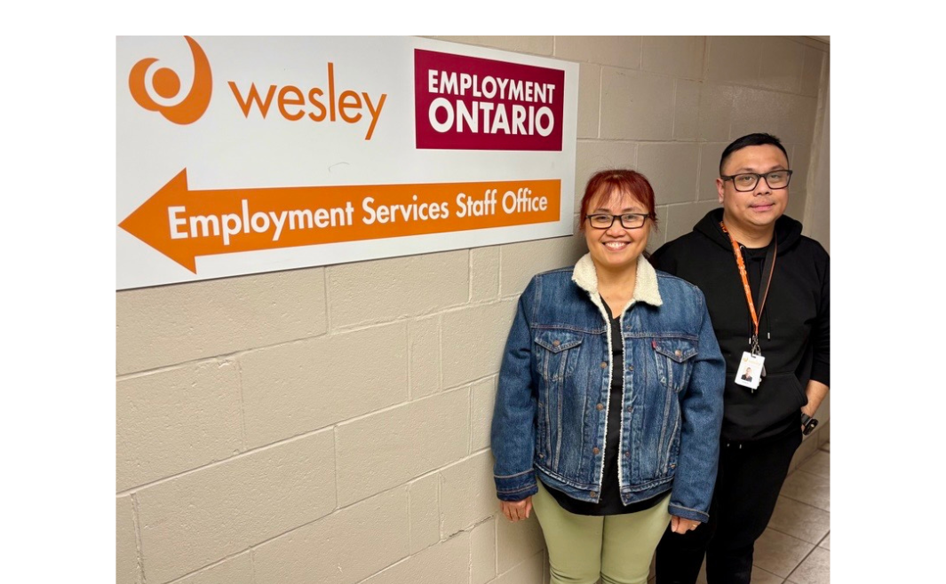 Two people stand smiling in a hallway beside a wall sign reading “Wesley Employment Ontario – Employment Services Staff Office,” with an arrow pointing left.
