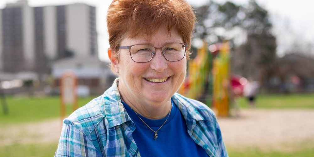 A woman with short reddish hair, glasses, and a blue shirt under a plaid shirt smiles outdoors in a park.