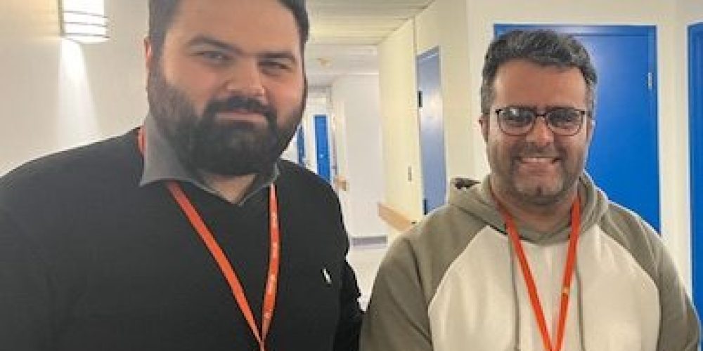 Two men stand side by side in a hallway, both wearing orange Wesley lanyards and looking at the camera, with blue doors visible in the background.