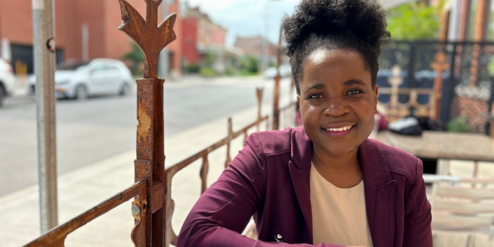 A woman in a maroon blazer sits at an outdoor cafe table, smiling at the camera with a city street in the background.