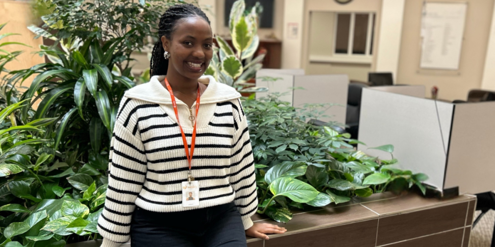 A woman wearing a striped sweater and ID badge smiles while standing next to indoor plants in an office setting.