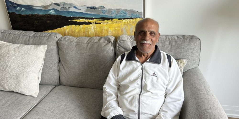 A senior Syrian man in a white Adidas jacket sits on a light gray couch, smiling.