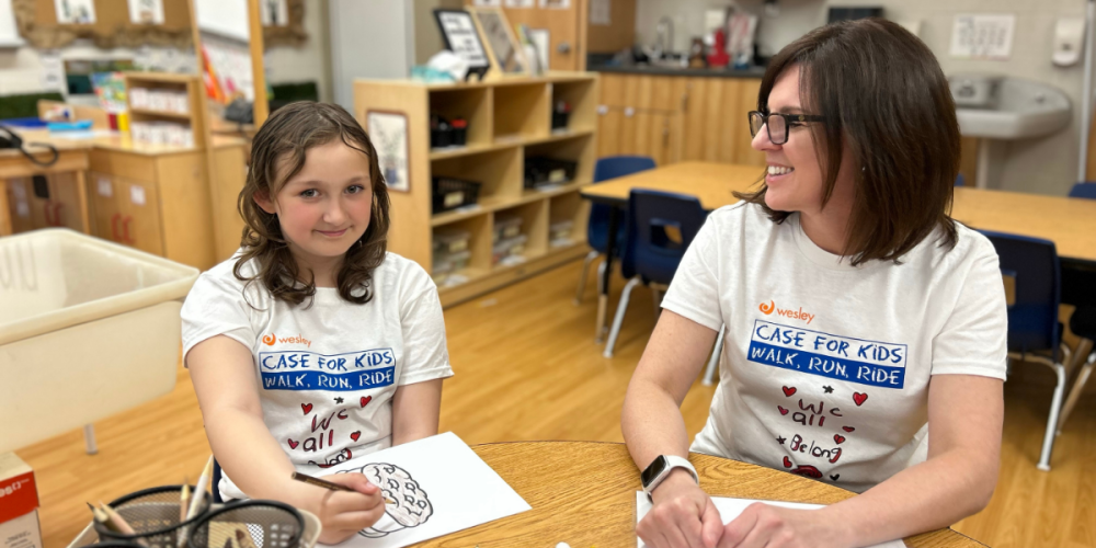 A young girl and a woman sit at a classroom table, smiling and drawing together. Both are wearing matching white 'Case for Kids: Walk, Run, Ride' t-shirts with the words 'We all belong' decorated with hearts.