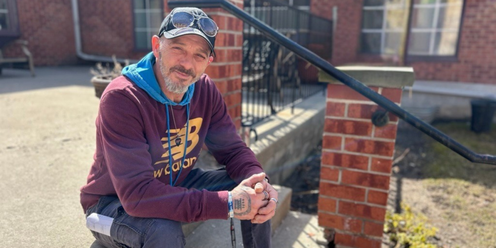 A man sits outside in the sun, wearing a burgundy hoodie, cap with sunglasses, and jeans. He has tattoos on his hands and a bandage on one finger. A brick building and railing are in the background.