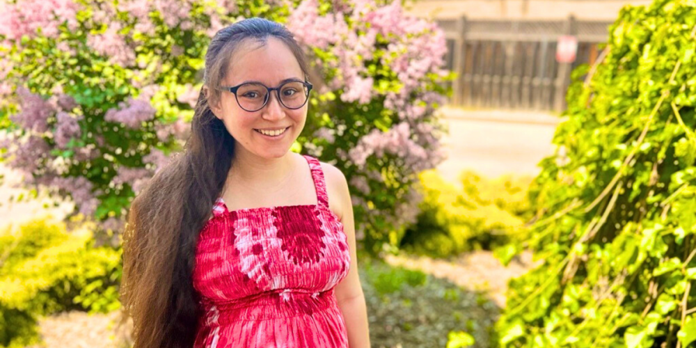 A woman with long brown hair and glasses stands outdoors, smiling at the camera. She is wearing a red and white tie-dye sleeveless dress. Behind her are blooming lilac bushes and other greenery on a sunny day.