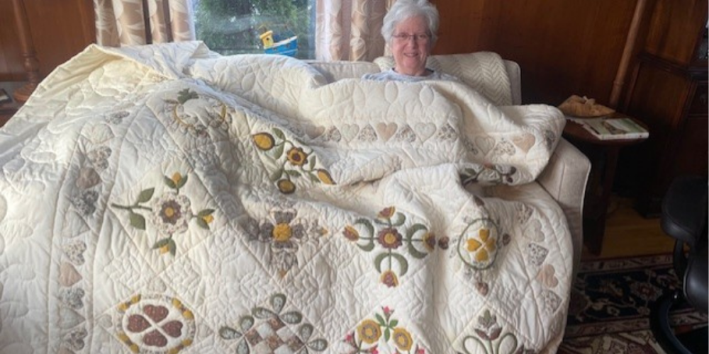 Woman smiling while sitting on a couch, covered with a large cream-colored quilt decorated with heart and flower patterns.