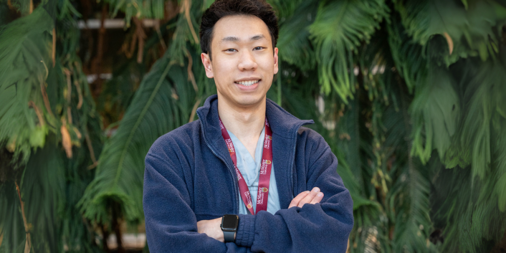 Kevin, wearing a blue fleece jacket and lanyard stands with arms crossed, smiling in front of large green leafy plants.