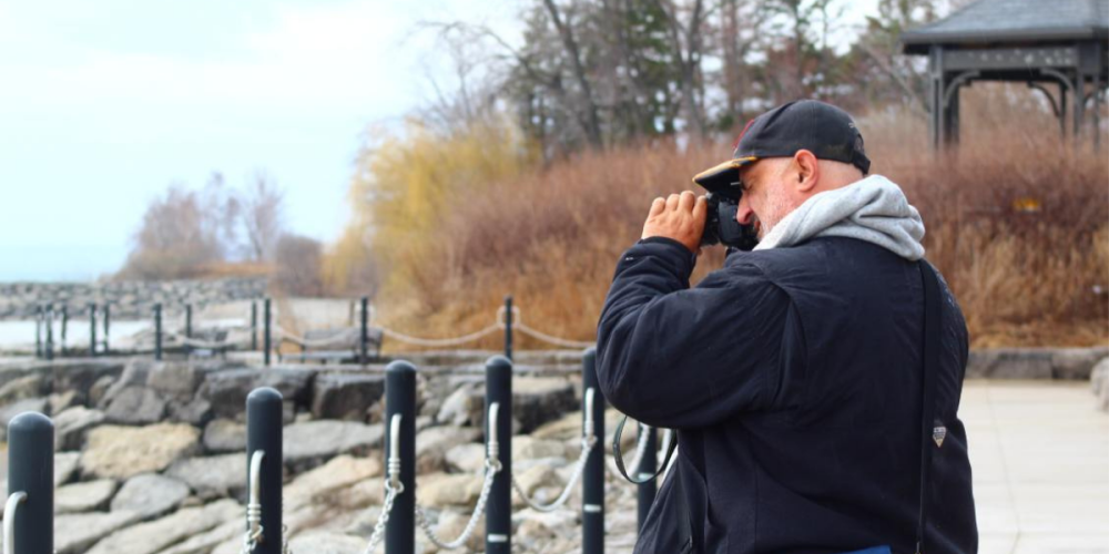 A man in a black jacket and cap stands on a paved path by the water, holding a camera to his face. Bare trees and rocks line the shore, with a wooden gazebo visible in the background.