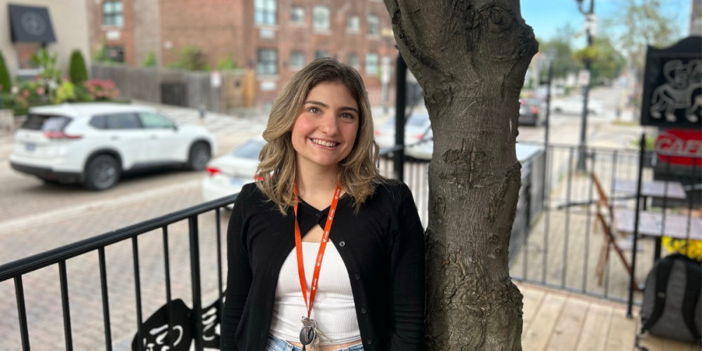 A young woman with a lanyard and keys stands next to a tree on an outdoor patio, smiling at the camera. Cars and brick buildings are visible in the background.