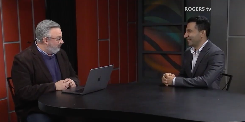 Two men sit across from each other at a desk during a Rogers TV interview. One man speaks while the other smiles and listens. A laptop sits on the desk between them in a television studio with red and dark-toned panels in the background.