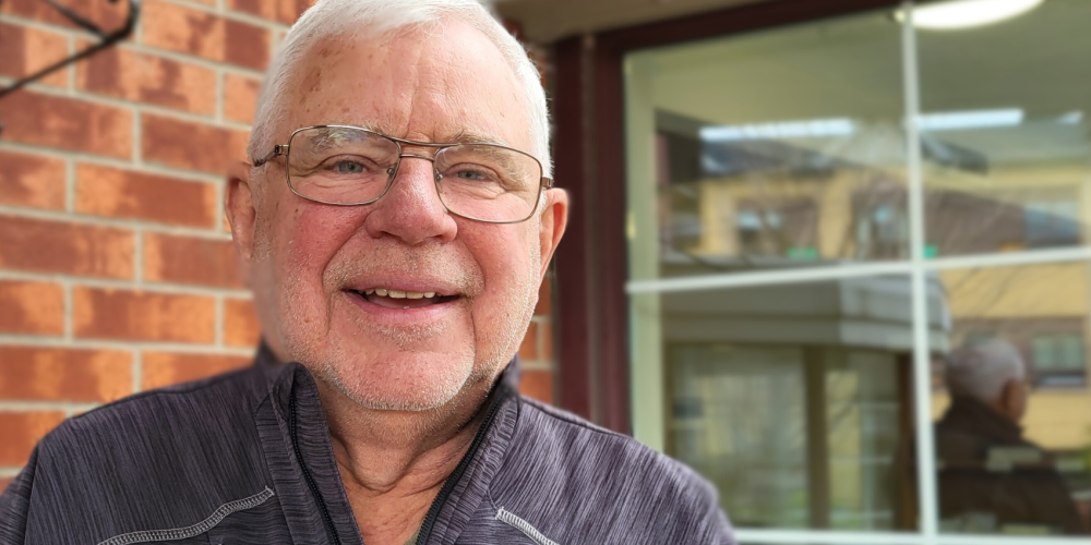 An older man with white hair, glasses, and a gray jacket smiles while standing in front of a brick wall and large window.