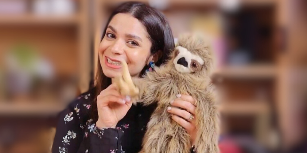 Dema, wearing a floral top smiles while holding a stuffed sloth.