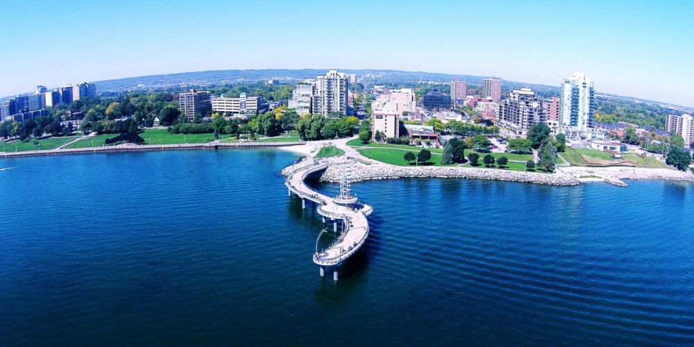 Aerial view of the Burlington waterfront with a curved pier extending into the blue water, surrounded by high-rise buildings and green spaces.