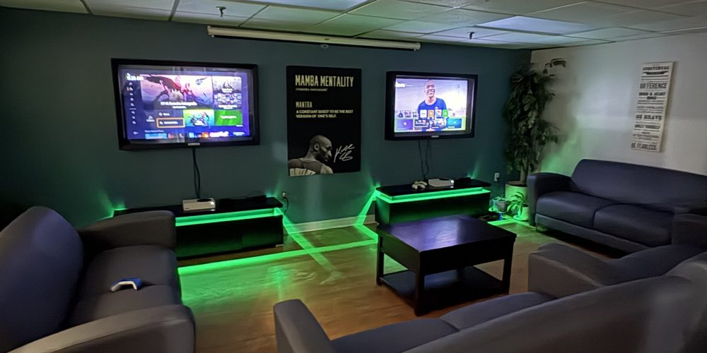 A cozy youth centre lounge with multiple blue couches arranged around a coffee table, two wall-mounted TVs displaying a video game menu, and gaming consoles below. Green LED accent lighting glows beneath the TV stands, creating a welcoming, modern space for youth to relax, play games, and socialize.