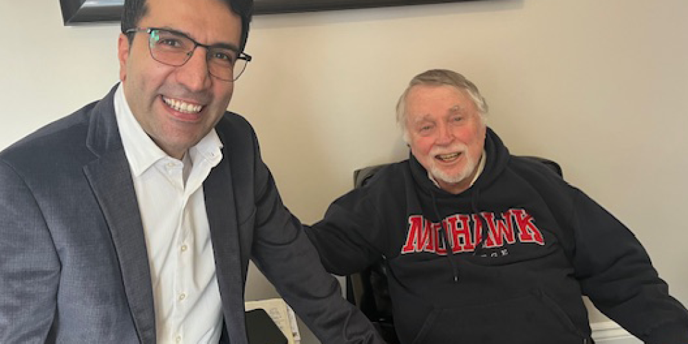 Two men are seated indoors, smiling at the camera. One wears a suit and glasses, the other wears a black hoodie.