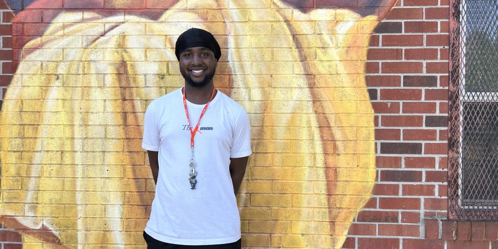 A person wearing a white t-shirt and black shorts stands smiling in front of a brick wall with a colourful mural.