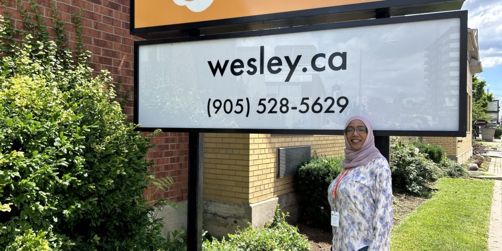Woman wearing a hijab and glasses stands outdoors in front of the Wesley main office sign.