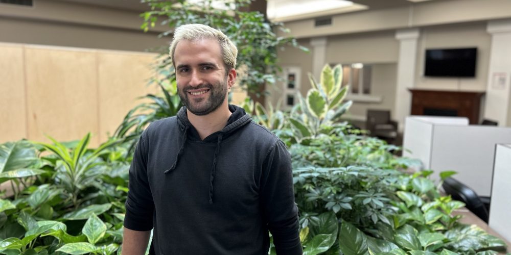 A man with short blonde hair and a dark beard, wearing a black hoodie, stands in front of indoor plants in an office setting.