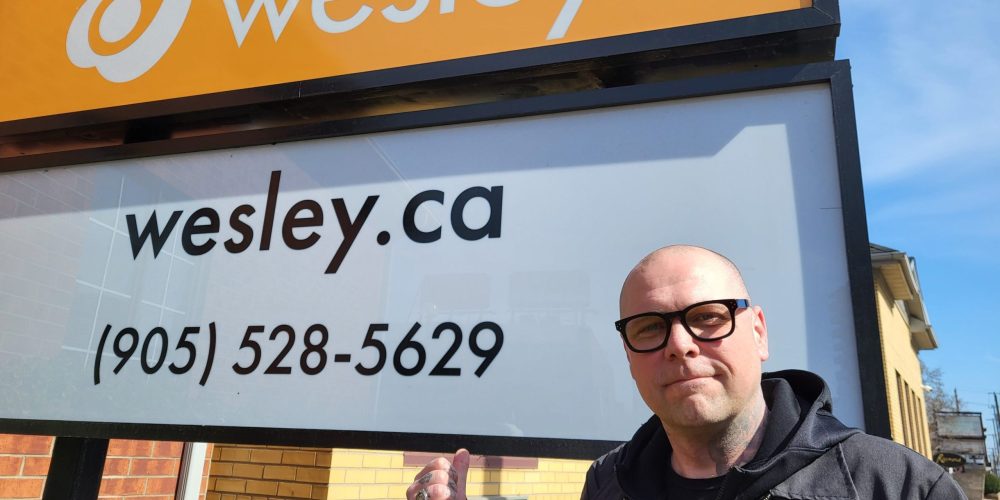 Joe, wearing glasses and a black jacket, stands outdoors, pointing at Wesley's main office sign.