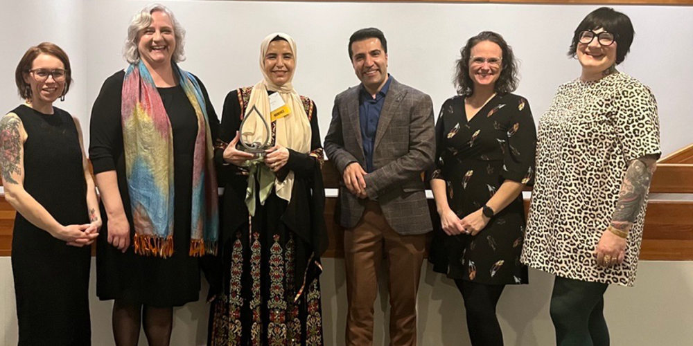 Six people stand side by side indoors, smiling at the camera. Lina is in the center, wearing a hijab and holding an award.