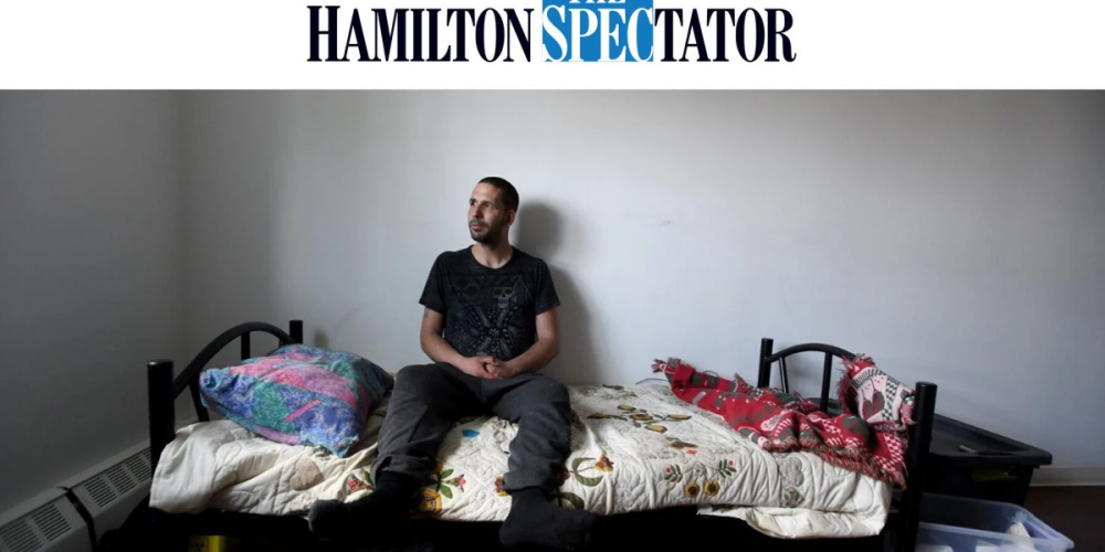 A man sits on the edge of a single bed in a sparsely furnished room, looking slightly to the side. The room has plain white walls and minimal decor, with a colorful pillow and quilt on the bed. The photo is from The Hamilton Spectator.