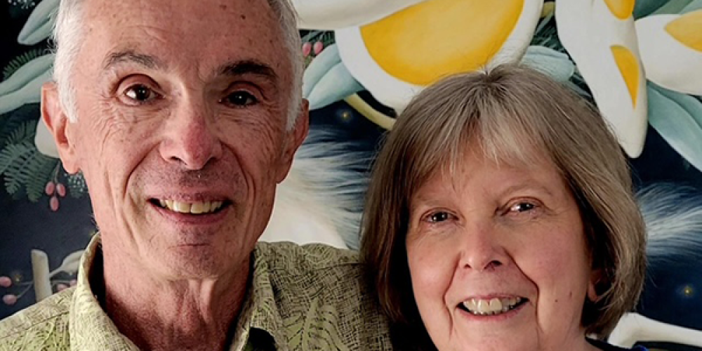 An older man and woman stand close together, smiling in front of a colorful painting.