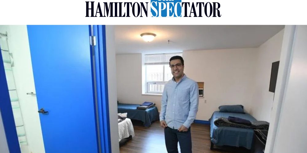 A man stands in a clean, simply furnished room with two single beds and blue doors, as seen in The Hamilton Spectator.