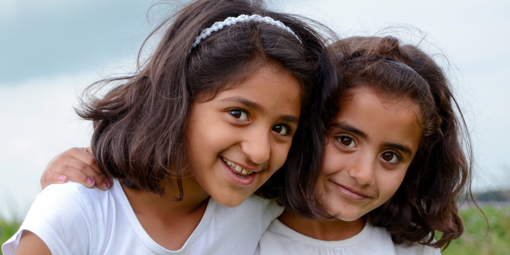 Two young girls with dark hair and white shirts stand close together outdoors, smiling at the camera.