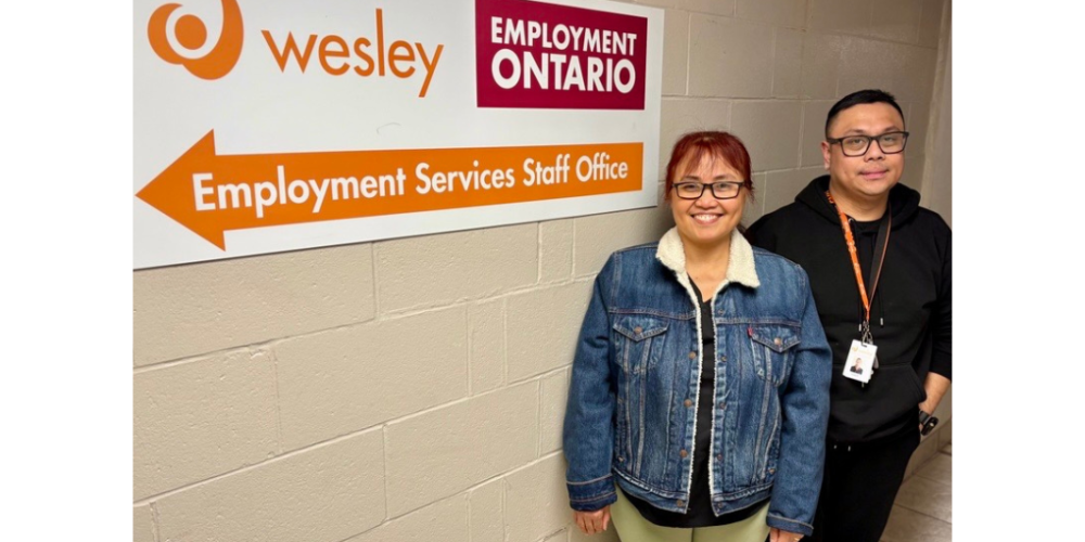 Two people stand smiling in a hallway beside a wall sign reading “Wesley Employment Ontario – Employment Services Staff Office,” with an arrow pointing left.