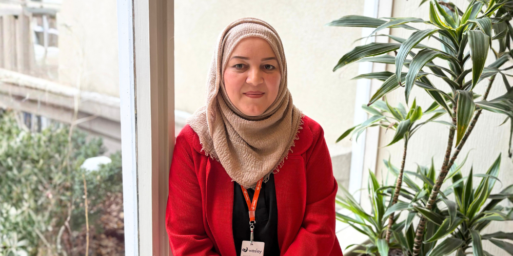 A woman wearing a beige hijab and red jacket sits by a bright window indoors, smiling gently at the camera. She has a Wesley staff lanyard around her neck, and green plants are visible beside her, with soft natural light filling the space.