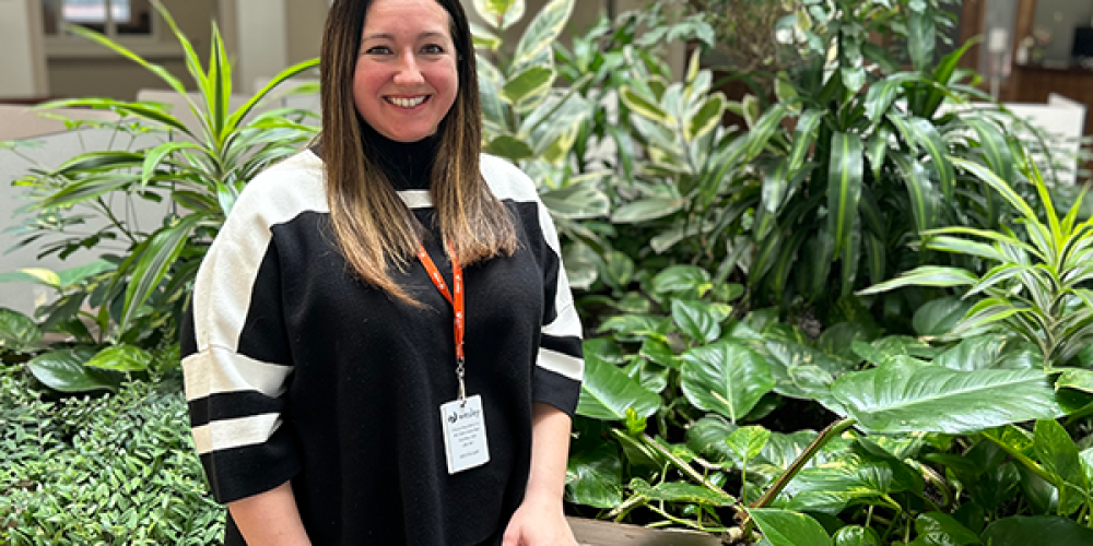 Keira, with long brown hair, wearing a black and white striped sweater and a Wesley name badge, stands and smiles in front of green plants indoors.